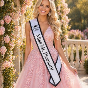 May include: A woman wearing a pink, embellished gown, a crown, and a sash that reads "Miss Mt. Pleasant" and "Queen 2026." She is smiling, with long blonde hair and earrings. The background features a floral archway.