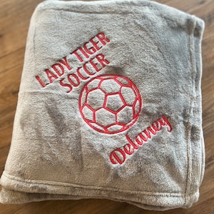 May include: A gray, plush blanket embroidered with "LADY TIGER SOCCER", a soccer ball, and the name "Delaney" in red thread. The blanket is folded and appears soft and cozy.