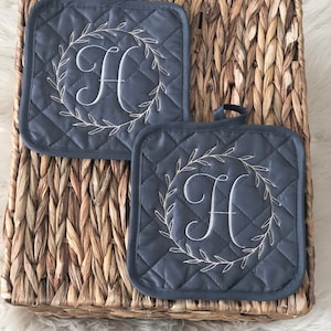 May include: Two gray quilted pot holders with a white embroidered wreath and the letter "H" in the center.