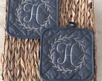 May include: Two gray quilted pot holders with a white embroidered wreath and the letter "H" in the center.