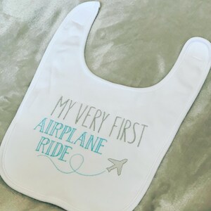 May include: White baby bib with the text "My Very First Airplane Ride" embroidered in gray and a blue airplane with a blue dotted line trail.