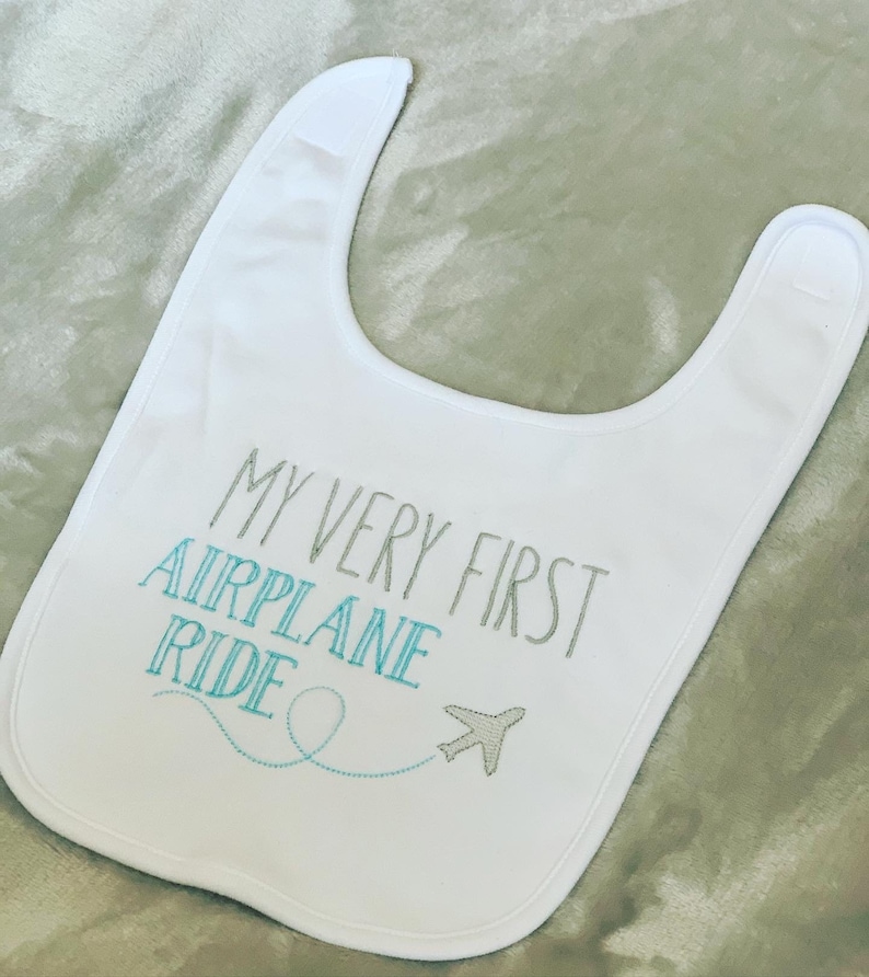 May include: White baby bib with the text "My Very First Airplane Ride" embroidered in gray and a blue airplane with a blue dotted line trail.