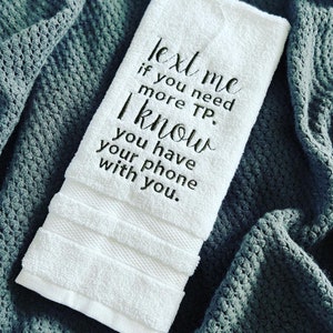 May include: White hand towel with black embroidered text that reads "Text me if you need more TP. I know you have your phone with you."