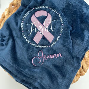 May include: A navy blue fleece blanket with a pink ribbon and the word "Fight" embroidered in the center. The ribbon is surrounded by a circle of words including "Hope", "Strength", "Courage", "Faith", "Family", "Love", "Support", "Awareness", "Relieve". The name "Joann" is embroidered below the ribbon.