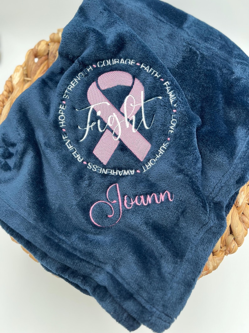 May include: A navy blue fleece blanket with a pink ribbon and the word "Fight" embroidered in the center. The ribbon is surrounded by a circle of words including "Hope", "Strength", "Courage", "Faith", "Family", "Love", "Support", "Awareness", "Relieve". The name "Joann" is embroidered below the ribbon.