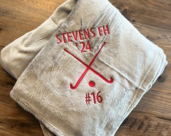 May include: A gray fleece blanket with red embroidered text that reads "Stevens FH '24 #16" and two crossed hockey sticks.