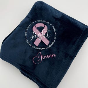 May include: A navy blue fleece blanket with a pink ribbon and the word "fight" embroidered in the center. The ribbon is surrounded by a circle of words including "hope", "strength", "courage", "faith", "family", "love", "support", "awareness", "rehab". The name "Joann" is embroidered below the ribbon.
