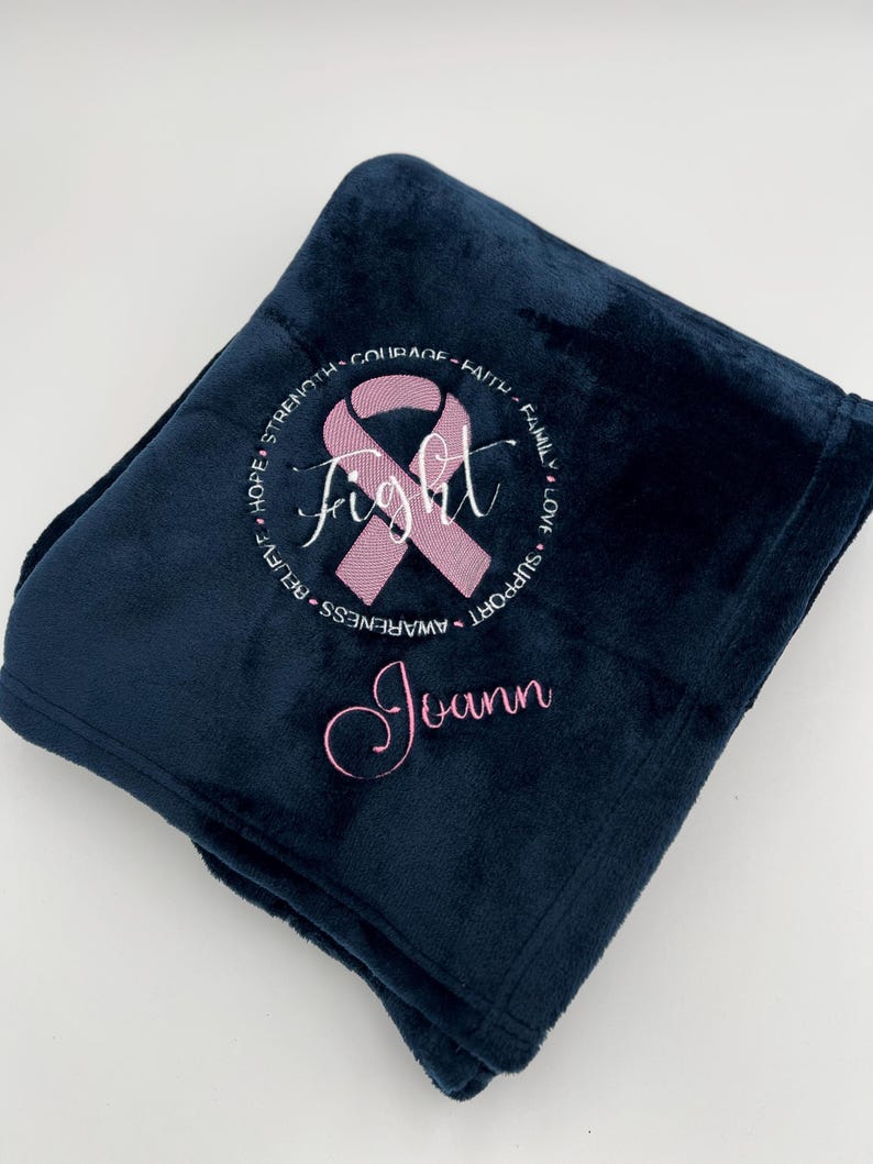 May include: A navy blue fleece blanket with a pink ribbon and the word "fight" embroidered in the center. The ribbon is surrounded by a circle of words including "hope", "strength", "courage", "faith", "family", "love", "support", "awareness", "rehab". The name "Joann" is embroidered below the ribbon.
