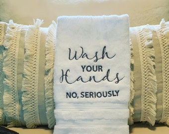 May include: White hand towel with the embroidered text "Wash Your Hands No, Seriously" on a bed with decorative pillows.