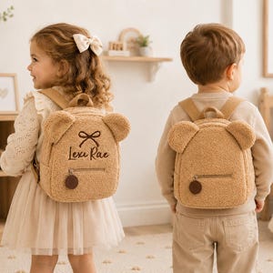 Puede incluir: Dos ni&ntilde;os con mochilas de osito de peluche marrones a juego. Las mochilas tienen orejas de oso, un lazo y el nombre "Lexi Rae" bordado en una. Las mochilas tienen cierre de cremallera y un charm redondo marr&oacute;n.