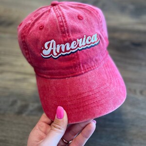Puede incluir: Una gorra de b&eacute;isbol roja con un "America" bordado en blanco en una fuente retro.
