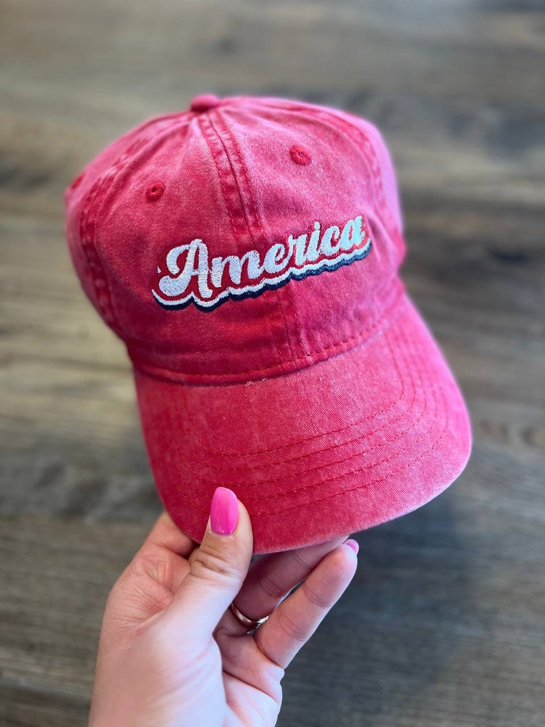 Puede incluir: Una gorra de b&eacute;isbol roja con un "America" bordado en blanco en una fuente retro.