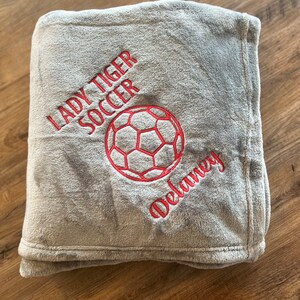 May include: A gray plush blanket with red embroidered text and a soccer ball design. The text reads "LADY TIGER SOCCER" and "Delaney". The blanket is folded and appears soft and cozy.