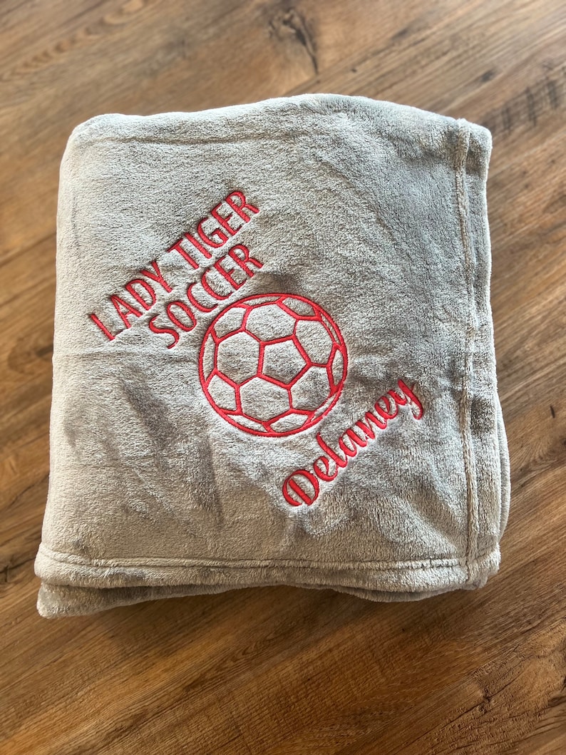May include: A gray plush blanket with red embroidered text and a soccer ball design. The text reads "LADY TIGER SOCCER" and "Delaney". The blanket is folded and appears soft and cozy.
