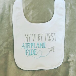 May include: White baby bib with the text "My Very First Airplane Ride" embroidered in gray and blue thread. A small airplane with a blue tail is embroidered next to the text.