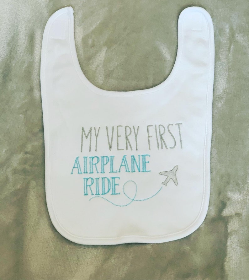 May include: White baby bib with the text "My Very First Airplane Ride" embroidered in gray and blue thread. A small airplane with a blue tail is embroidered next to the text.