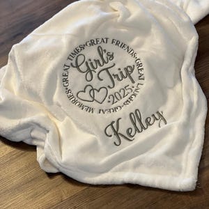 May include: White plush blanket with embroidered text "Girls Trip 2025" in a circle with hearts and the name "Kelley" below.