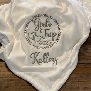 May include: White plush blanket with embroidered text "Girl's Trip 2025" in a circle with hearts and the text "Great Times, Great Friends, Great Laughs, Great Memories". The name "Kelley" is embroidered below the circle.