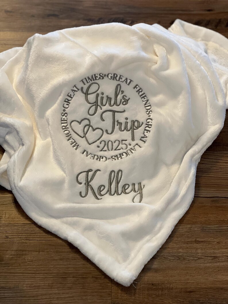 May include: White plush blanket with embroidered text "Girl's Trip 2025" in a circle with hearts and the text "Great Times, Great Friends, Great Laughs, Great Memories". The name "Kelley" is embroidered below the circle.