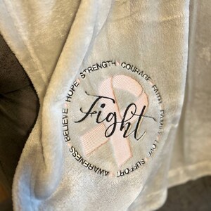 May include: A soft, gray fleece blanket with a pink ribbon and the word "fight" embroidered in black. The ribbon is surrounded by the words "believe, hope, strength, courage, faith, family, love, support, awareness".