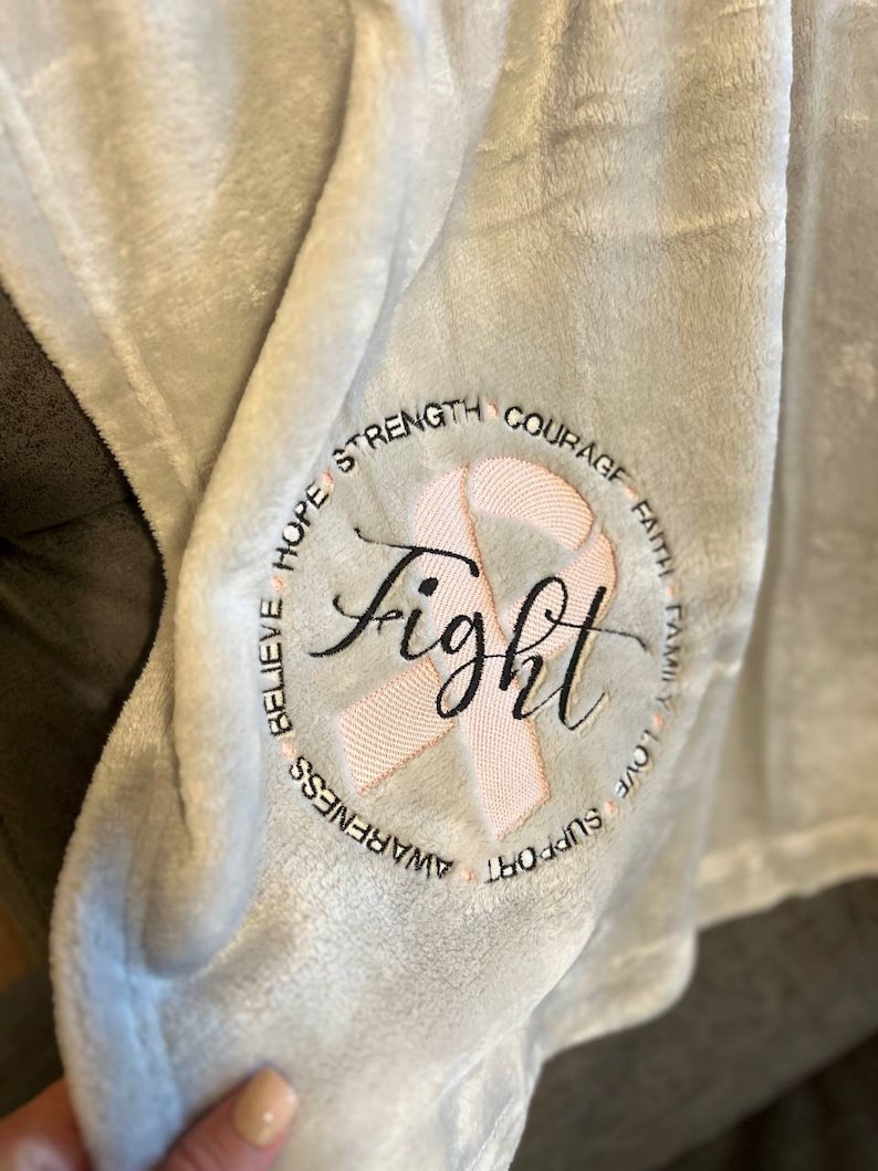 May include: A soft, gray fleece blanket with a pink ribbon and the word "fight" embroidered in black. The ribbon is surrounded by the words "believe, hope, strength, courage, faith, family, love, support, awareness".