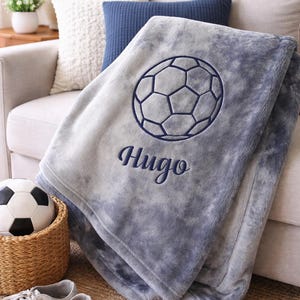May include: A gray and blue soccer-themed blanket with the name "Hugo" embroidered in navy blue. The blanket features a soccer ball design and is folded over a sofa. A soccer ball and shoes are in the foreground.