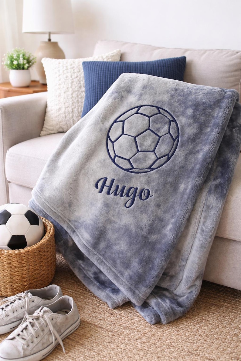 May include: A gray and blue soccer-themed blanket with the name "Hugo" embroidered in navy blue. The blanket features a soccer ball design and is folded over a sofa. A soccer ball and shoes are in the foreground.