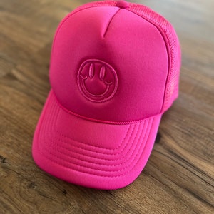 K&ouml;nnte beinhalten: Leuchtend pinke Trucker-Cap mit einem Smiley-Emblem. Die Kappe hat eine Mesh-R&uuml;ckseite und einen gebogenen Schirm. Das Smiley-Gesicht ist auf der Vorderseite aufgestickt.