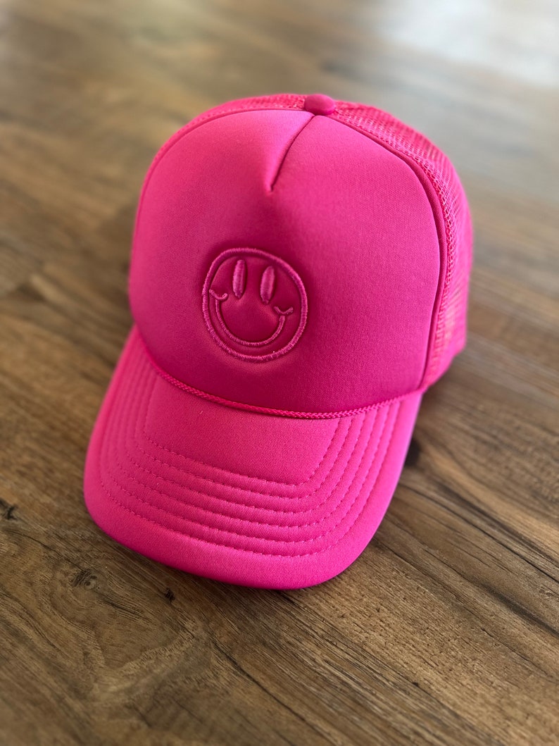K&ouml;nnte beinhalten: Leuchtend pinke Trucker-Cap mit einem Smiley-Emblem. Die Kappe hat eine Mesh-R&uuml;ckseite und einen gebogenen Schirm. Das Smiley-Gesicht ist auf der Vorderseite aufgestickt.