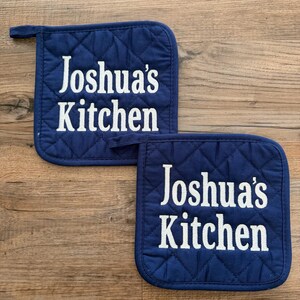 May include: Two navy blue quilted pot holders with white embroidered text that reads "Joshua's Kitchen". The pot holders have a diamond pattern and a small loop for hanging. They are placed on a wooden surface.