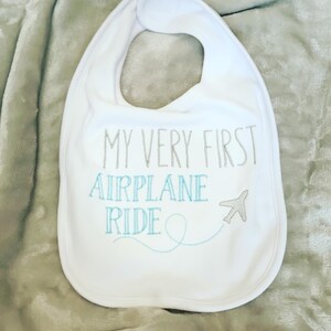 May include: White baby bib with the text "My Very First Airplane Ride" embroidered in gray and blue thread. A small airplane icon is embroidered next to the text.