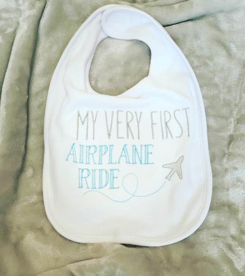 May include: White baby bib with the text "My Very First Airplane Ride" embroidered in gray and blue thread. A small airplane icon is embroidered next to the text.