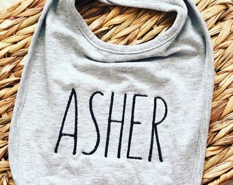 May include: Gray cotton baby bib with a black embroidered name "Asher" on it.