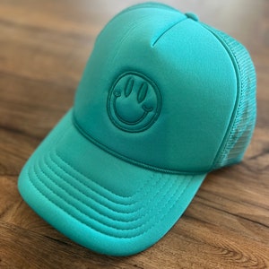 K&ouml;nnte beinhalten: T&uuml;rkisfarbene Trucker-M&uuml;tze mit Smiley-Aufn&auml;her. Die M&uuml;tze hat einen gebogenen Schirm, eine Mesh-R&uuml;ckseite und einen aufgestickten Smiley. Die M&uuml;tze ist aus weichem Material und liegt auf einer Holzoberfl&auml;che.