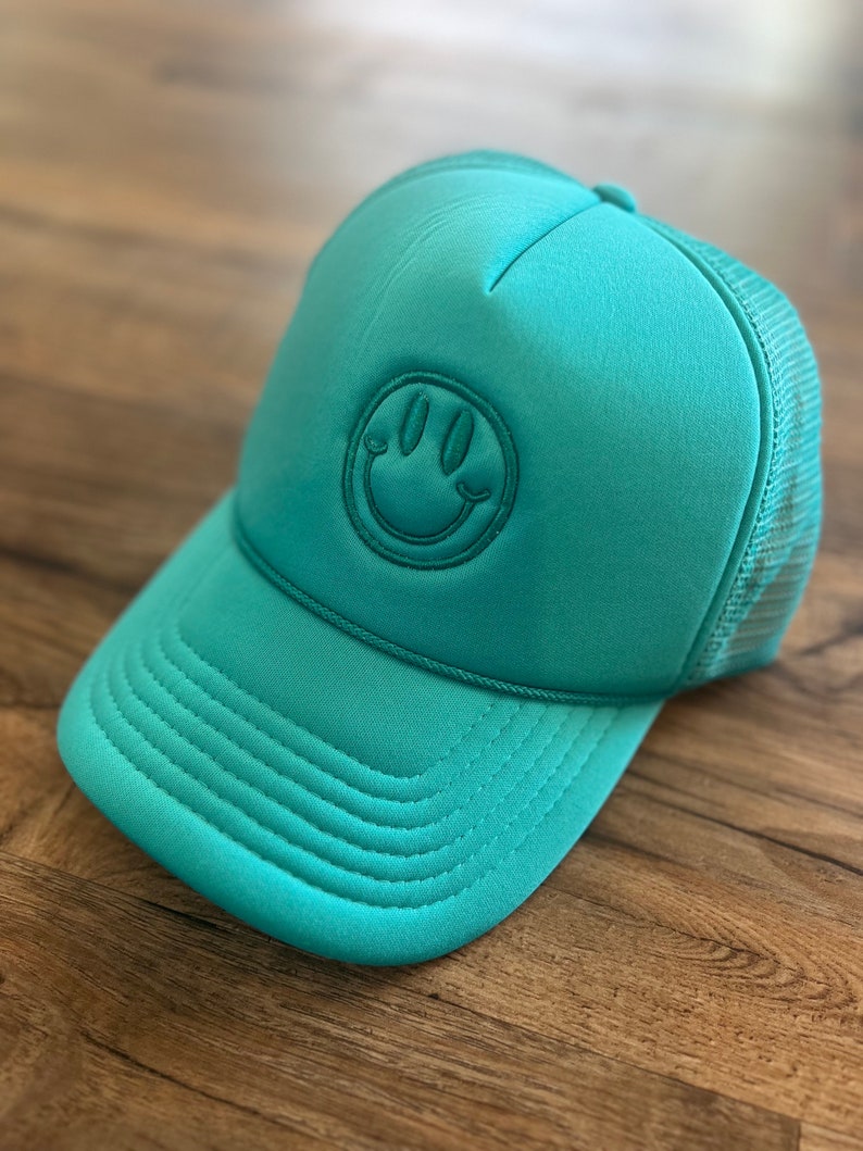 K&ouml;nnte beinhalten: T&uuml;rkisfarbene Trucker-M&uuml;tze mit Smiley-Aufn&auml;her. Die M&uuml;tze hat einen gebogenen Schirm, eine Mesh-R&uuml;ckseite und einen aufgestickten Smiley. Die M&uuml;tze ist aus weichem Material und liegt auf einer Holzoberfl&auml;che.