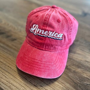 Puede incluir: Una gorra de b&eacute;isbol roja con la palabra "America" bordada en la parte delantera en blanco con detalles azules.