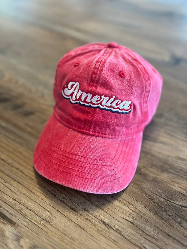 Puede incluir: Una gorra de b&eacute;isbol roja con la palabra "America" bordada en la parte delantera en blanco con detalles azules.