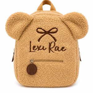 Puede incluir: Una mochila en forma de oso de peluche de color marr&oacute;n claro con un peque&ntilde;o bolsillo frontal y un asa superior. La mochila tiene dos grandes protuberancias en forma de oreja y un detalle de lazo marr&oacute;n. El nombre "Lexi Rae" est&aacute; bordado en escritura marr&oacute;n. Una etiqueta circular marr&oacute;n con una imagen de oso cuelga de la cremallera.