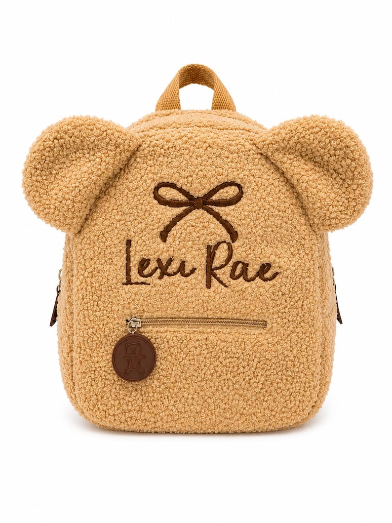 Puede incluir: Una mochila en forma de oso de peluche de color marr&oacute;n claro con un peque&ntilde;o bolsillo frontal y un asa superior. La mochila tiene dos grandes protuberancias en forma de oreja y un detalle de lazo marr&oacute;n. El nombre "Lexi Rae" est&aacute; bordado en escritura marr&oacute;n. Una etiqueta circular marr&oacute;n con una imagen de oso cuelga de la cremallera.