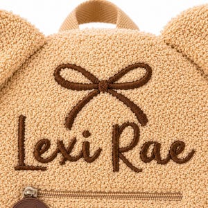 Puede incluir: Una mochila en forma de oso de peluche de color beige con el nombre "Lexi Rae" bordado en escritura marr&oacute;n. La mochila tiene un detalle de lazo marr&oacute;n y un peque&ntilde;o bolsillo con cremallera en la parte inferior. Las orejas son una caracter&iacute;stica prominente.