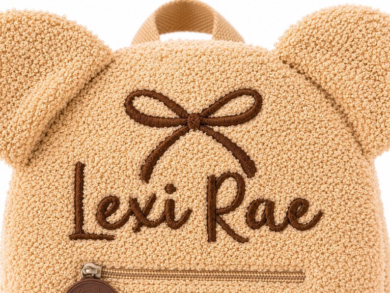 Puede incluir: Una mochila en forma de oso de peluche de color beige con el nombre "Lexi Rae" bordado en escritura marr&oacute;n. La mochila tiene un detalle de lazo marr&oacute;n y un peque&ntilde;o bolsillo con cremallera en la parte inferior. Las orejas son una caracter&iacute;stica prominente.