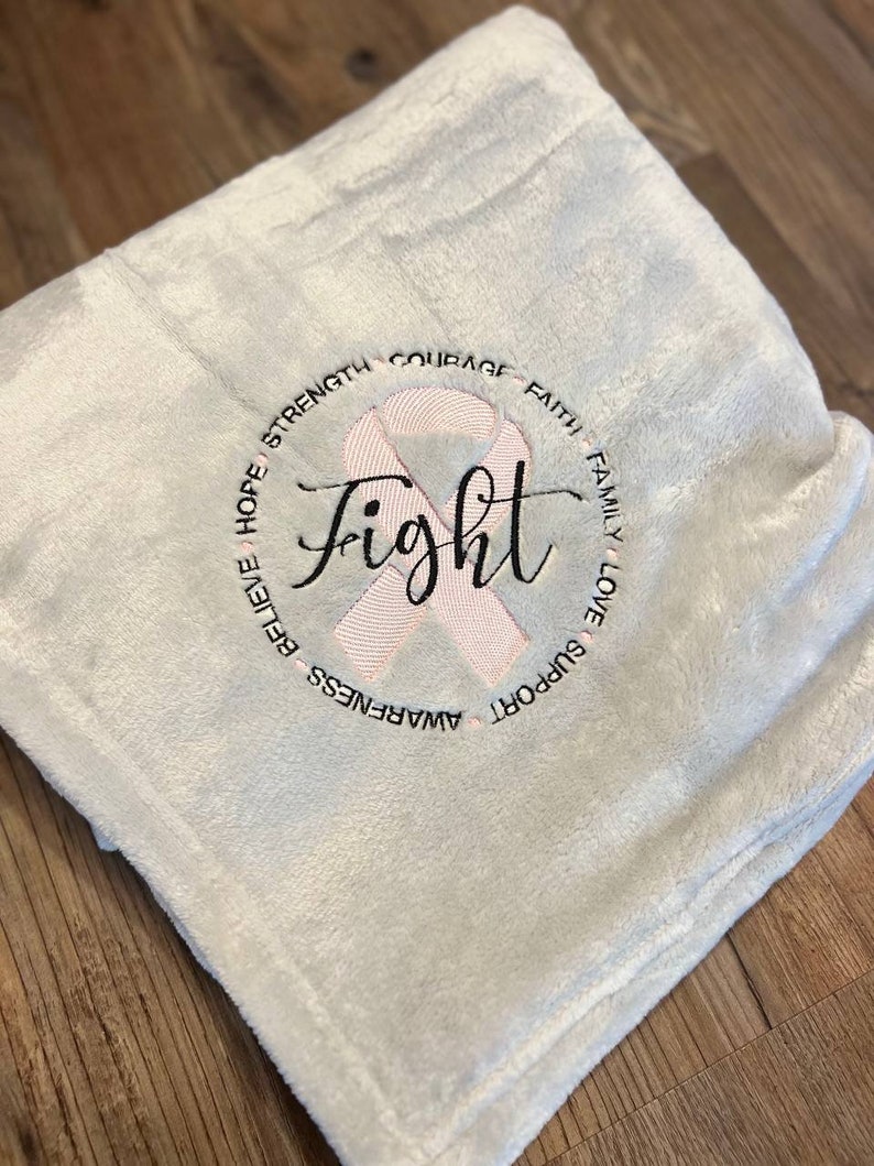 May include: A soft, white fleece blanket with a pink ribbon and the word "fight" embroidered in a circle. The circle also includes the words "hope, strength, courage, faith, family, love, support, awareness, believe".