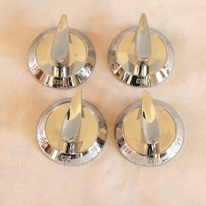 May include: Four vintage stove knobs with a silver and gold finish. Each knob has a silver handle and a circular base with markings. The knobs are arranged in a square formation against a neutral background.