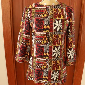 May include: A long-sleeved tunic featuring a patchwork-style print in shades of red, black, yellow, and white. The garment displays a repeating pattern of geometric shapes, stylized floral motifs, and abstract lines across the entire fabric surface.