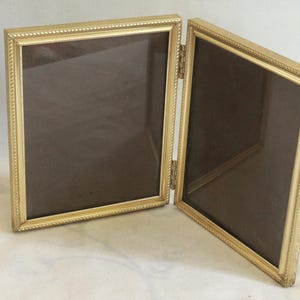 May include: A double-hinged picture frame with a metallic gold finish and a textured, beaded border design. The two rectangular frames are connected by a central metal hinge, allowing the unit to stand upright in an open, angled position.
