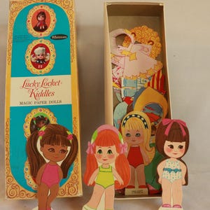 May include: Vintage "Lucky Locket Kiddles" Magic Paper Dolls set. The box is teal and yellow with illustrations of the dolls. Several paper dolls are standing in front of the box, with various outfits and hairstyles. The box is open, revealing more paper doll clothing.