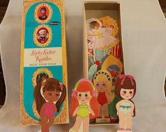 Pode incluir: Conjunto de bonecas de papel mágicas "Lucky Locket Kiddles" vintage. A caixa é turquesa e amarela com ilustrações das bonecas. Várias bonecas de papel estão em frente à caixa, com várias roupas e penteados. A caixa está aberta, revelando mais roupas de bonecas de papel.