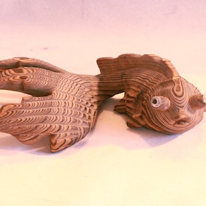 May include: A wooden sculpture of a fish with a detailed, layered design. The fish has large, bulging eyes and a flowing tail. The wood grain is visible, creating a natural, textured appearance. The sculpture is a decorative item.