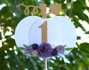 Op de afbeelding: Witte pompoen cake topper met een gouden glitter nummer één en paarse papieren bloemen.
