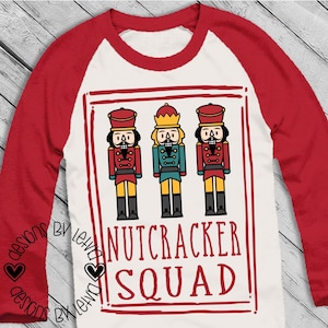 May include: White long-sleeve shirt with red sleeves. The shirt has a graphic of three nutcracker soldiers in a red frame with the text "Nutcracker Squad!" in red.